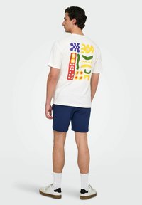 Camiseta blanca de manga corta con impresiones abstractas coloridas en púrpura, rojo, verde y amarillo en la parte posterior. Combinada con pantalones cortos azul marino y zapatillas.