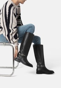 Stivali neri in pelle fino al ginocchio con dettagli in argento, caratterizzati da una punta arrotondata e una texture liscia, indossati con jeans blu.