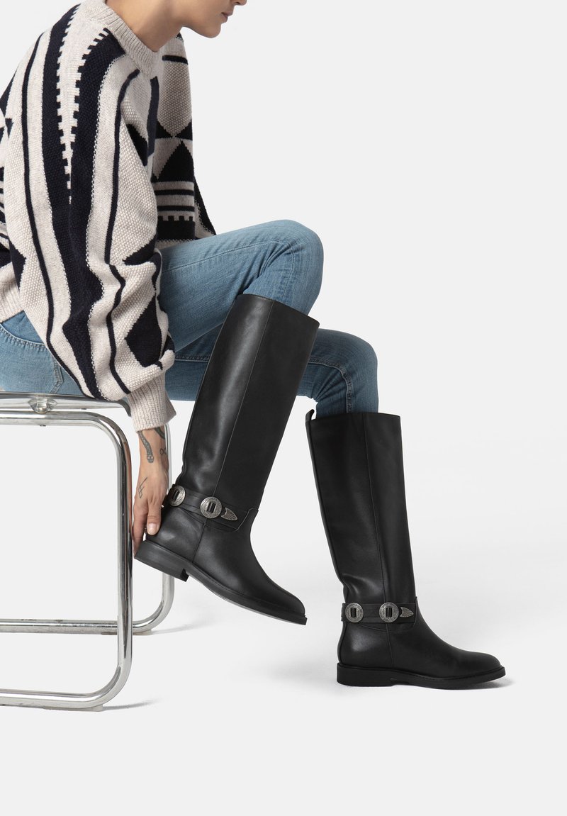 Stivali neri in pelle fino al ginocchio con dettagli in argento, caratterizzati da una punta arrotondata e una texture liscia, indossati con jeans blu.