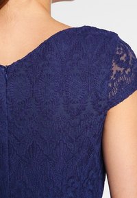 Námorně modré krajkové šaty s kulatým výstřihem a krátkými rukávy. Obsahují složité květinové vzory a jemnou strukturu, s detailem na zip na zadní části.
