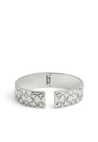 Silverfärgad manschettarmband med ingraverade cirkulära mönster och små nitar. Har en gångjärnsstängning för enkel användning. Slät yta, polerad finish.