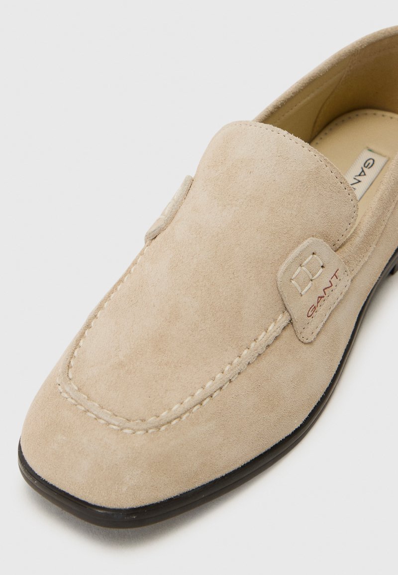 Scarpa slip-on in suede beige con punta tondeggiante, cuciture decorative e un piccolo logo stampato sul lato. Suola nera a profilo basso.