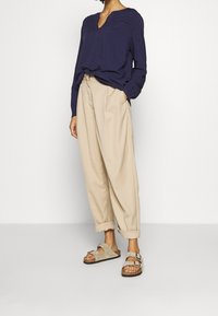 Blouse bleu marine à manches longues avec détails texturés et col en V, associée à un pantalon beige ample et des sandales beige clair à deux boucles.