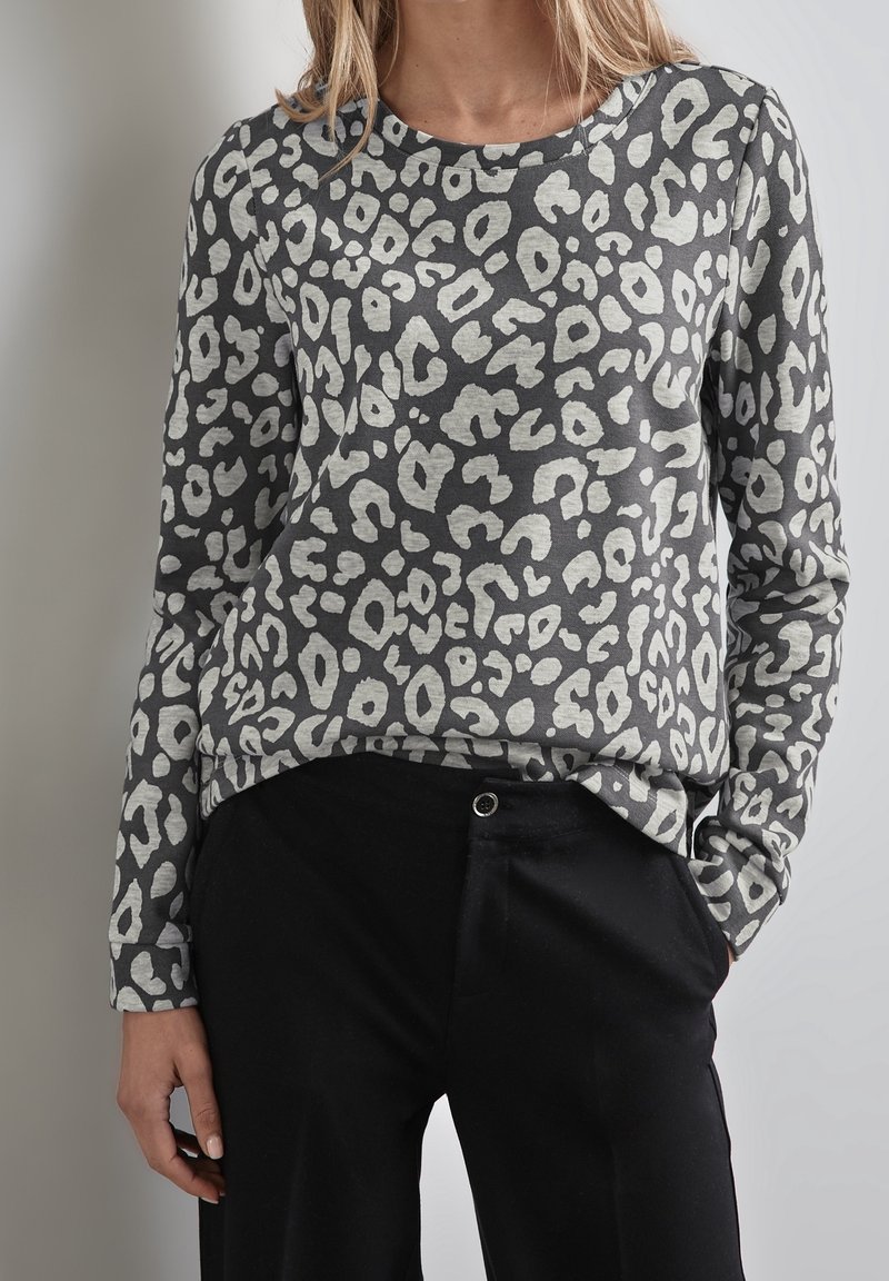 Femme portant un haut gris à manches longues avec imprimé léopard blanc, associée à un pantalon noir, debout devant un fond clair uni.