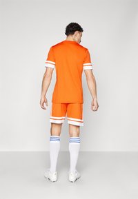 Camiseta deportiva naranja con rayas blancas en las mangas, pantalones cortos naranjas a juego, calcetas blancas hasta la rodilla con rayas azules y zapatillas blancas.