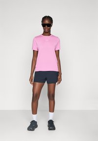 Roze sportska majica kratkih rukava, uparena s tamnim šortsevima. Model nosi crne sunčane naočale i tamne tenisice s bijelim čarapama.