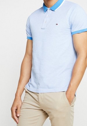 Mann trägt hellblaues kurzärmeliges Poloshirt mit blauem Kragen und beiger Hose, eine Hand in der Tasche vor weißem Hintergrund.