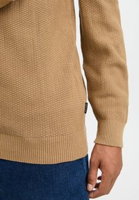 Strickpullover in Hellbraun mit einem strukturierten Muster, gerippten Bündchen und Saum, versehen mit einem kleinen Markenetikett an der Seite.