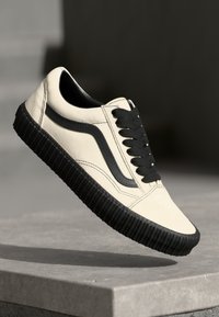 Zapatilla de lona beige con suela de goma negra y acentos. Presenta cordones negros y un diseño de franja lateral. Textura suave y silueta de caña baja.