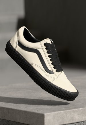 Beige canvas sneaker with black rubber sole and accents. Features black laces and a side stripe design. Smooth texture and low-top silhouette.
