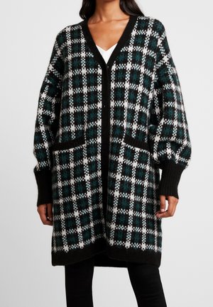 Nainen pukeutuneena pitkään, ylisuurikokoiseen musta-valko-vihreäruudulliseen neuletakkiin, jossa etutaskut ja musta reunus, mustujen housujen ja valkoisen paidan päällä.