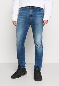Slim-fit blue denim jeans with a faded finish, standard five-pocket design, and belt loops, paired with black athletic shoes.