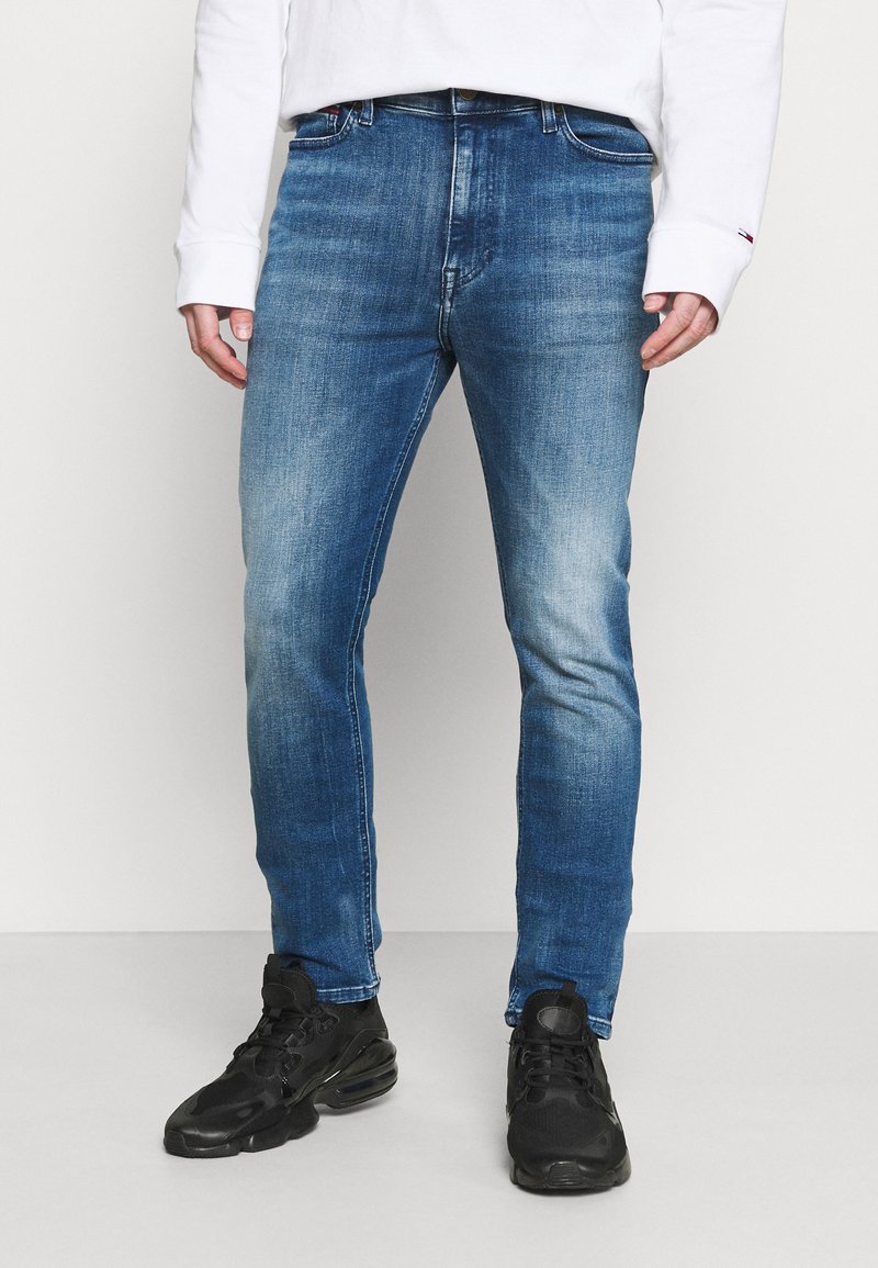 Slim-fit blue denim jeans with a faded finish, standard five-pocket design, and belt loops, paired with black athletic shoes.