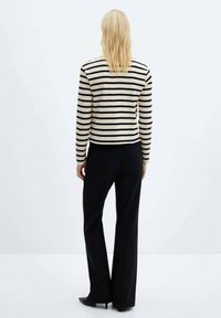 Gestreifter Pullover in Schwarz und Beige, im cropped Design, kombiniert mit schwarzen weit geschnittenen Hosen und hohen Schuhen, vor einem schlichten Hintergrund.
