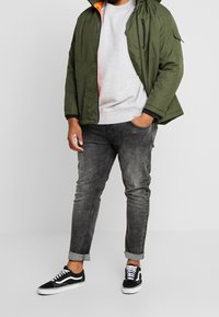Mann mit grüner Kapuzenjacke, hellgrauem Sweatshirt, hochgekrempelten ausgebleichten schwarzen Jeans und schwarz-weißen Sneakern, der mit einer Hand in der Tasche steht.