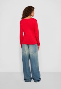 Vrouw met krullend haar, gekleed in een rode lange mouwen top en wijdvallende vergrijsde blauwe jeans, staand met het gezicht naar een effen lichtgrijze muur.