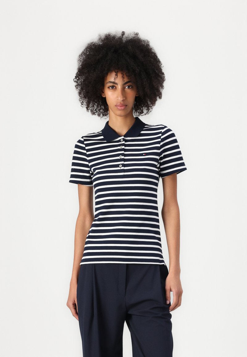 Femme aux cheveux bouclés portant un polo à manches courtes rayé bleu marine et blanc et un pantalon bleu marine, debout devant un fond blanc uni.