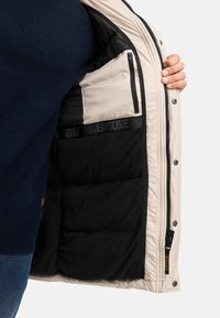 Beige jas met een zwarte fleece voering. Voorzien van een binnenzak met rits en een elastische band met merknaam. Buitenmateriaal is glad en gewatteerd.