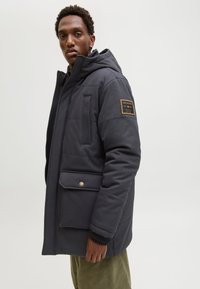 Ciemnoszara parka z kapturem o fakturowanym wykończeniu, z naszywką z logo na lewym rękawie, przednimi kieszeniami i zapięciem na zamek.