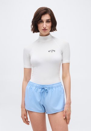 Γυναίκα με κοντά καστανά μαλλιά, φορούν λευκή κοντομάνικη μπλούζα για προστασία από τον ήλιο και ανοιχτό μπλε μαγιό με κορδόνι, στέκεται μπροστά σε απλό φόντο.