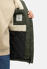 Zelená bunda s huňatým krémovým golierom a prešívanou čiernou podšívkou. Obsahuje vnútornú vrecko s etiketou. Viditeľný zips a patentky.