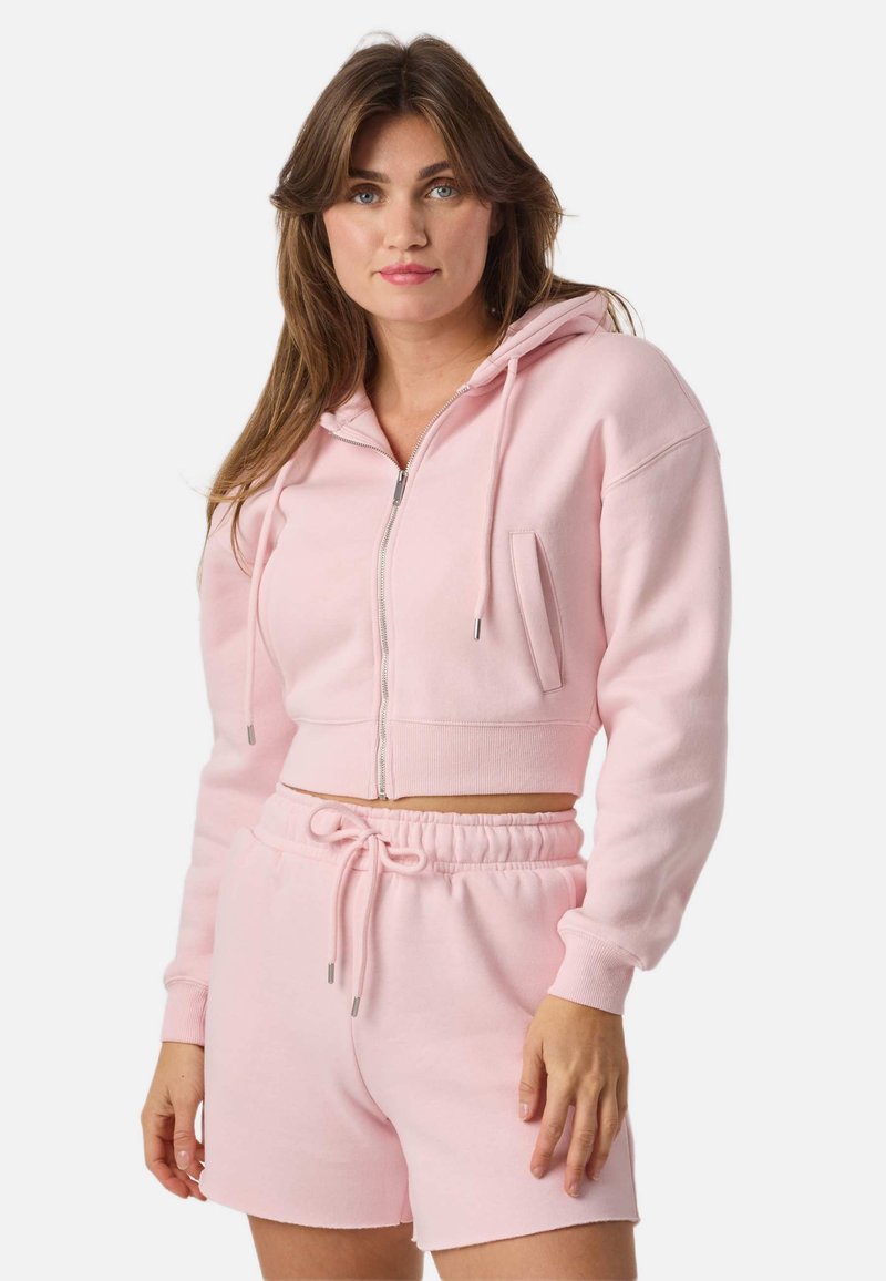 Femme portant un sweat à capuche zippé court rose clair et un short assorti à cordon de serrage, debout devant un fond blanc uni.