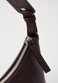 Sac en cuir bourgogne foncé présentant une surface texturée, une fermeture éclair argentée, et une sangle robuste avec un détail en métal au point de fixation.