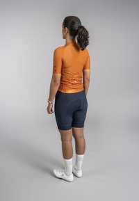 Oranje korte mouwen wielershirt van textuurstof, gecombineerd met marineblauwe gevoerde shorts. Witte fietsschoenen en gestreepte sokken maken de outfit compleet.