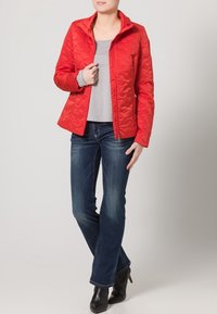 Röd vadderad jacka med hög krage, dragkedja och långa ärmar, tillsammans med en grå topp och mörkblå jeans. Svarta ankelstövlar.