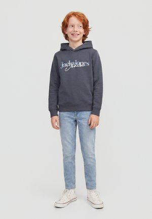 Rothaariger Junge trägt einen dunkelgrauen Hoodie mit Logo, hellblaue Jeans und weiße Sneakers und steht vor einem schlichten hellen Hintergrund.