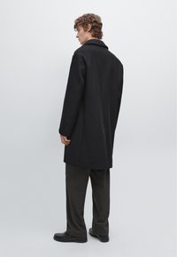 Zwarte wollen jas met een rechte snit, voorzien van een geplooide kraag en lange mouwen, gecombineerd met donker geruite broek. Schoenen zijn van zwart leer.