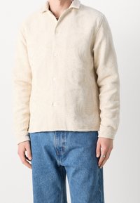 Krämfärgad skjorta med knapp-up-funktion som har ett strukturerat blommönster, långa ärmar och klassisk krage, matchad med blå denimbyxor.