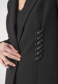 Černé sako s elegantním designem, které má krajkové detaily na rukávu, vyrobené z hladké látky a doplněné kovovými oky.
