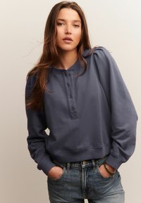 Vrouw met lang bruin haar, gekleed in een blauwe knopenhoodie en jeans, met handen in de zakken, staande tegen een effen lichte achtergrond.