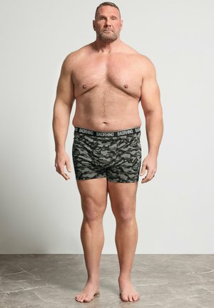 Uomo muscoloso di mezza età in piedi a piedi nudi su pavimento grigio, indossa boxer mimetici con fascia "BADRHINO" davanti a un muro bianco.