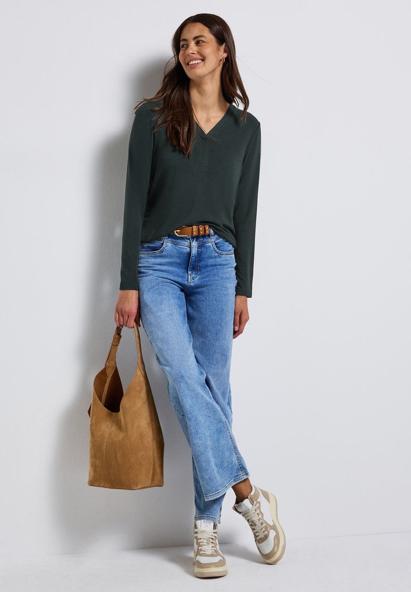 Haut long à manches vertes avec col en V, jeans droits bleu clair, sac fourre-tout en suède beige, et baskets blanches avec des accents beige.