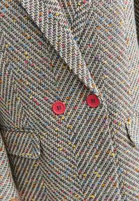 Sak na míru z texturované látky s herringbone vzorem a vícebarevnými flíčky. Obsahuje dva červené knoflíky a klopový límec.