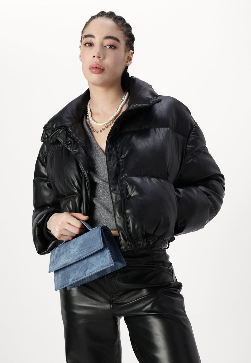 Schwarze Steppjacke mit hohem Kragen, glänzender Textur und Knopfdetails. Kombiniert mit einer blauen Wildleder-Clutch und grauem Oberteil, verziert mit geschichteten Perlen.