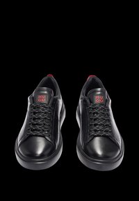 Baskets en cuir noir avec des lacets texturés, bout rond et semelle légèrement épaisse. Marquage rouge sur la languette. Finition lisse et design minimaliste.