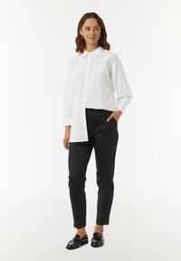 Weißes Hemd mit langen Ärmeln und leicht asymmetrischem Design. Kombiniert mit schwarzen, enganliegenden Hosen und glänzenden schwarzen Slippern.