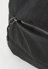 Mustekkan musta reppu, jossa on vetskarilla suljettava tasku, tekstuurinen pinta, metallinen vetoketjun vetopidike ja näkyvät ompeleet. Kompakti ja minimalistinen muotoilu.