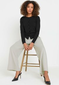 Schwarze Langarm-Cardigan mit Knopfverschluss, getragen mit hellgrauen, weiten Hosen und schwarzen Slingback-Pumps, sitzt auf einem Holzstuhl.