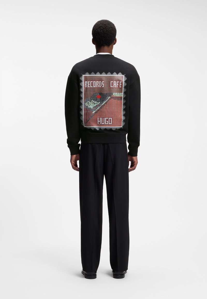 Homme debout de dos, portant un sweat-shirt noir avec un dessin d’un tourne-disque et le texte "RECORDS CAFE HUGO" dans le dos.