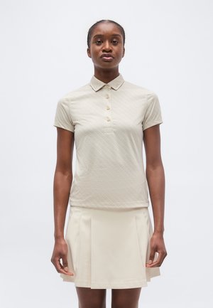 Mujer con un polo beige de manga corta y una falda plisada color crema, de pie contra un fondo claro y liso.