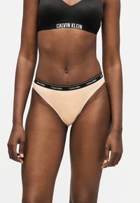 Femme portant un soutien-gorge de sport noir Calvin Klein et une culotte beige avec une ceinture noire, les deux arborant le logo Calvin Klein.