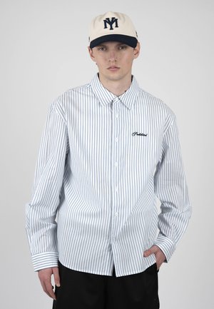 Junger Mann trägt ein weißes, fein gestreiftes Hemd mit „Political“-Stickerei, beige Kappe mit marineblauem Logo und schwarze Hose, steht mit einer Hand in der Tasche.