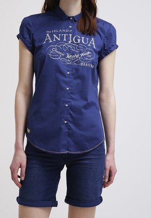 Femme portant une chemise bleu marine à manches courtes avec le texte "Antigua Classic Yacht Week" et un short en jean bleu foncé retroussé.