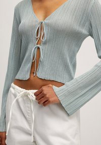 Helles blaues geripptes Crop-Top mit V-Ausschnitt und Schleifenverschluss, mit ausgestellten Ärmeln und einem weichen, strukturierten Stoff. Kombiniert mit weißen Hosen.