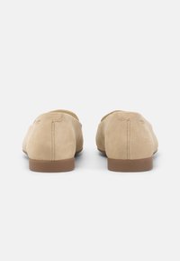 Zapatos de ante beige sin cordones con suela plana marrón. Textura suave, diseño minimalista y costuras visibles en el talón.