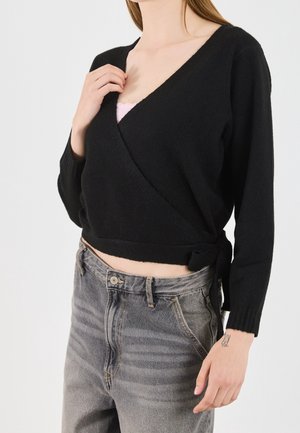 Kvinde iført en sort omslagsstriktrøje bundet i taljen, grå højtaljede jeans og en synlig lyserød camisole under.
